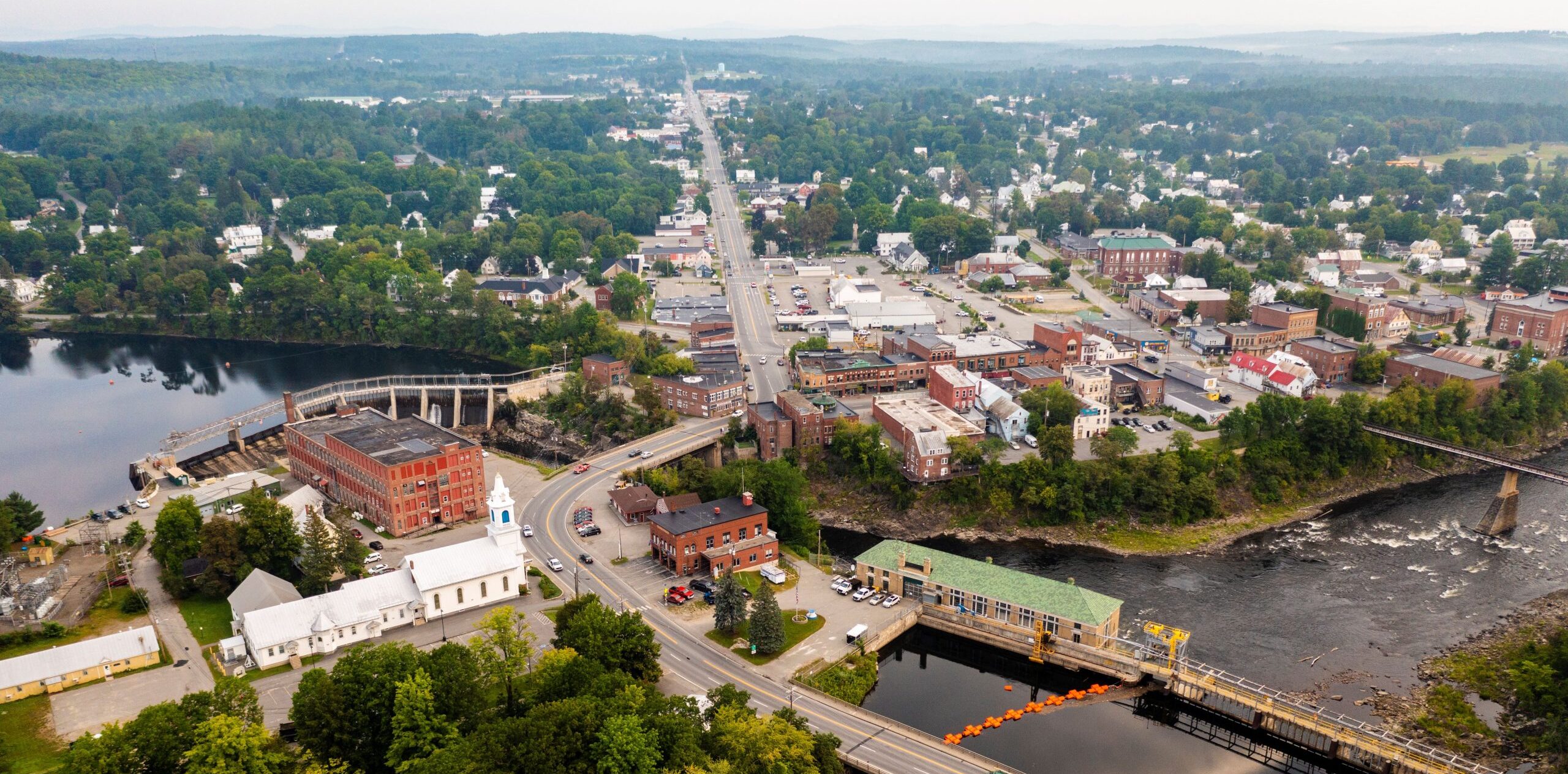 Aerial view of a riverside town with tree-lined neighborhoods, bridges, a dam, and surrounding hills