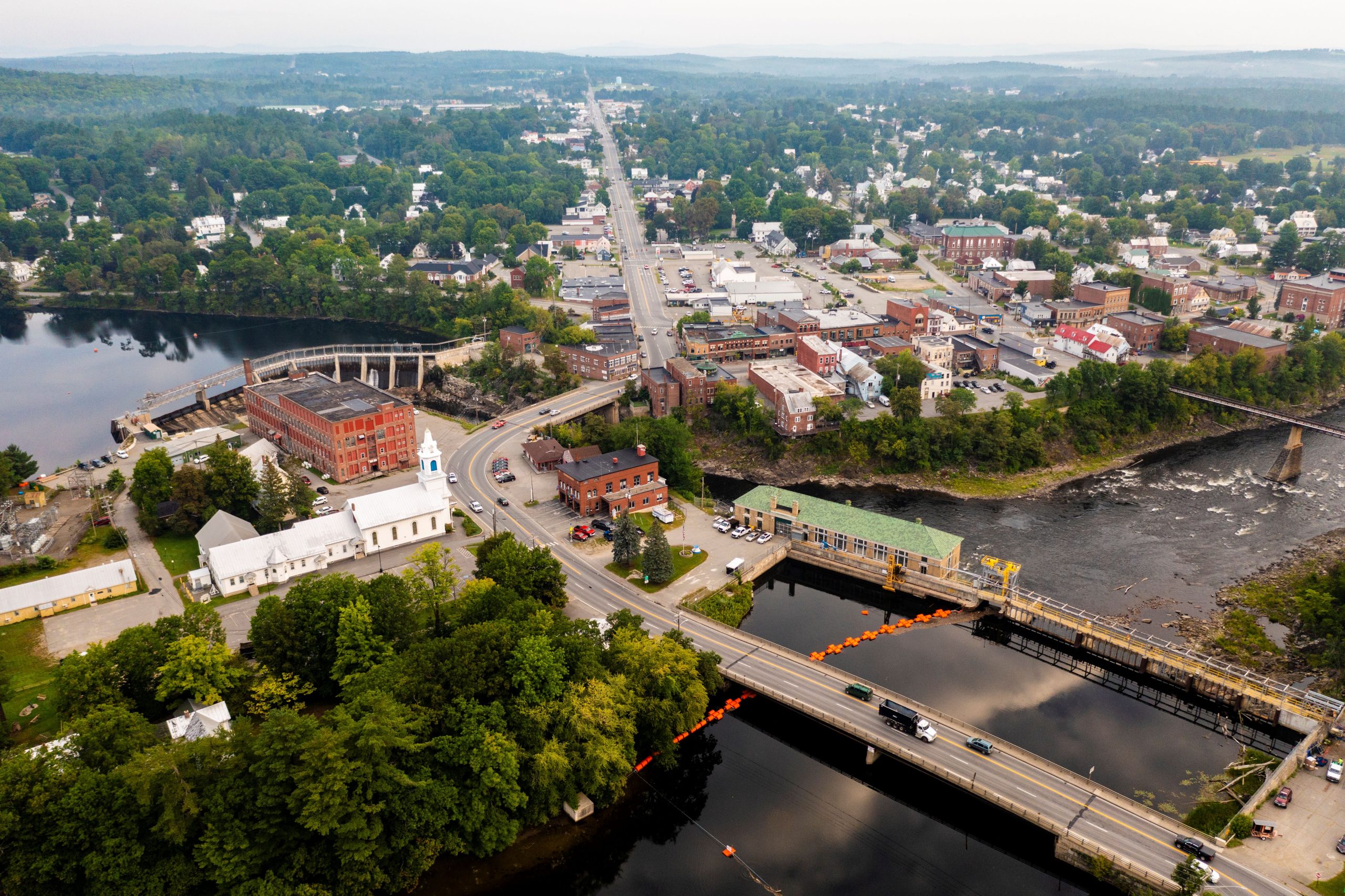 Aerial view of a riverside town with tree-lined neighborhoods, bridges, a dam, and surrounding hills