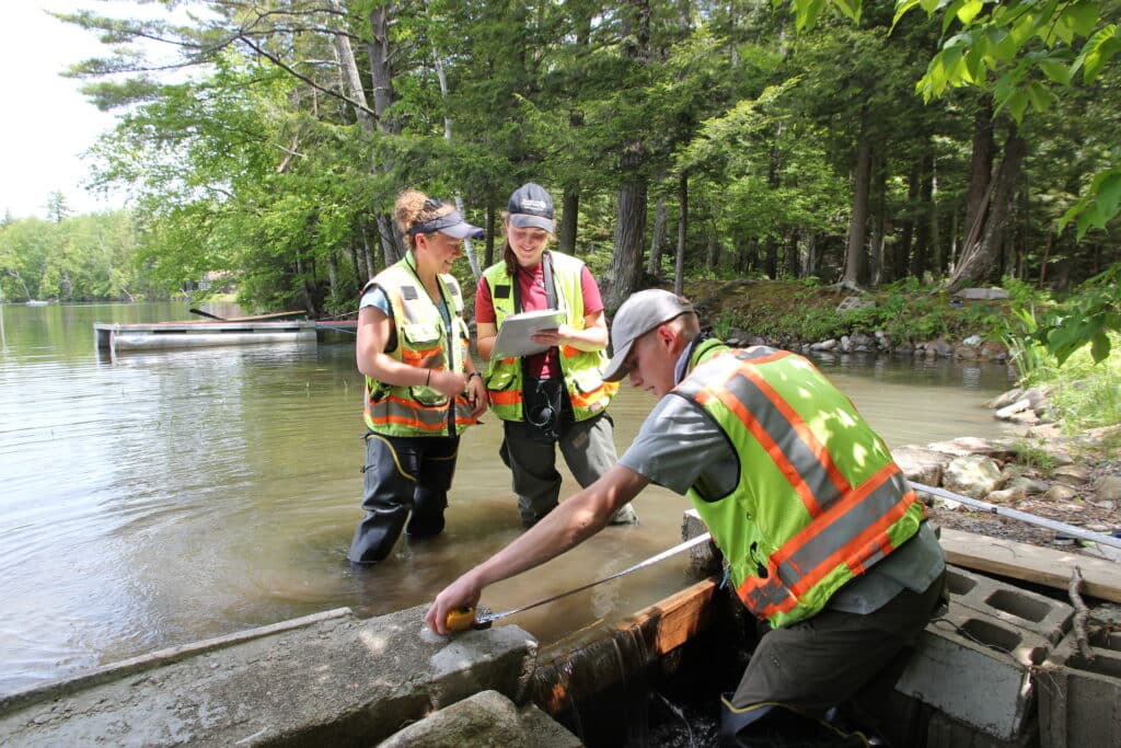 Three environmental workers measure a concrete structure at lake near a forested shoreline, with one recording data on a tablet