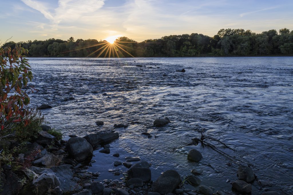 Sunset over a rocky river bordered by trees and shoreline vegetation