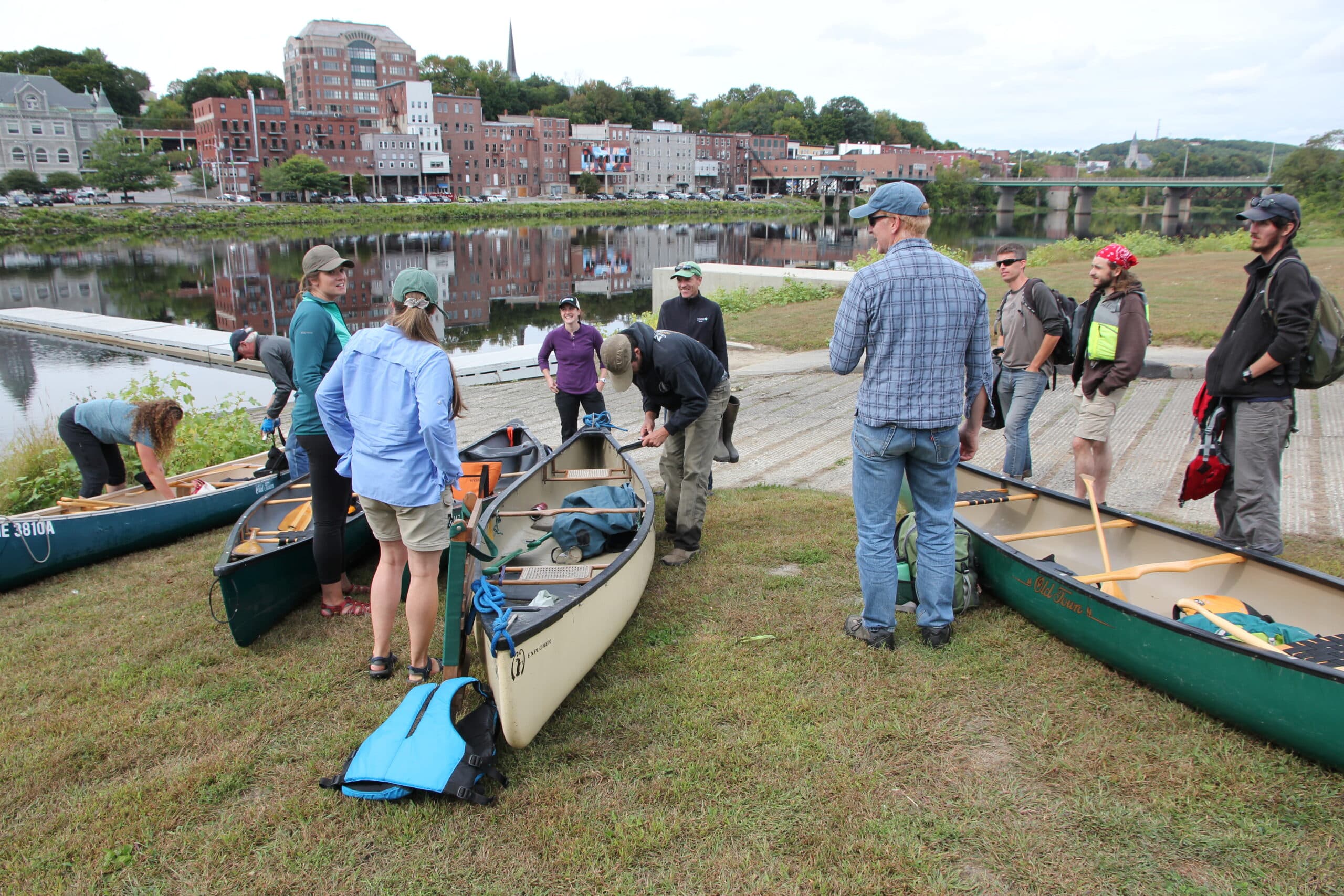 People prepare canoes beside a river, with a town and bridge across the water